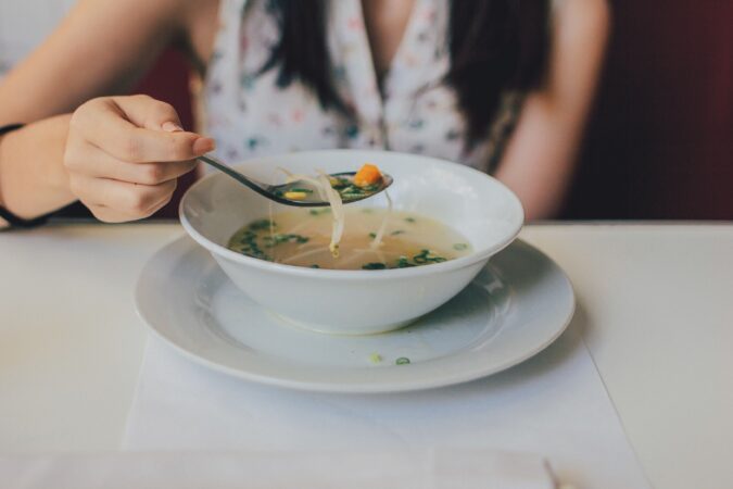 Spoonful of soup lifted from white bowl on white plate with partial torso of woman in background.