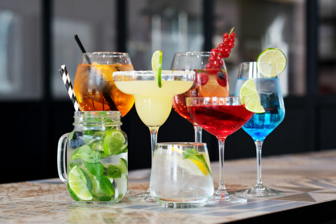 Seven cocktails in various shaped glasses sitting on bar.
