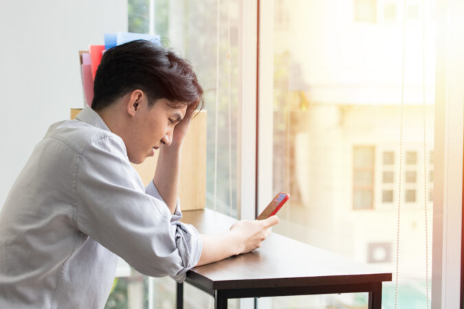 Young Asian man sitting at desk in front of window with left hand on his forehead looking at phone in his right hand. He looks frustrated.