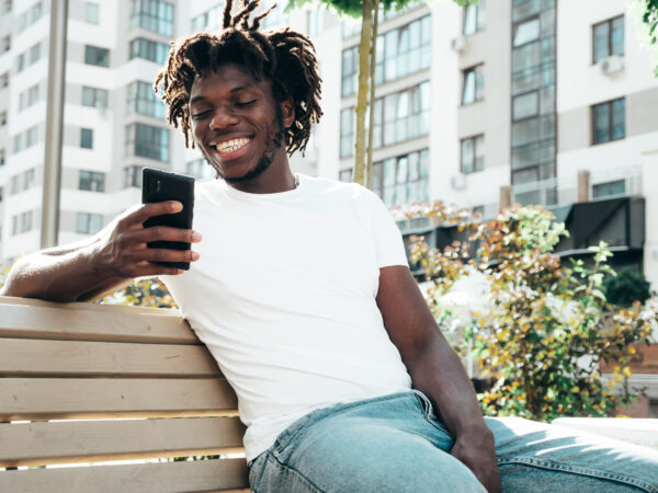 African American man sitting on bench smiling while searching on phone.