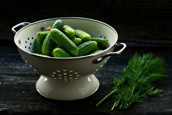 Porcelain colander filled with cucumbers sitting on dark wood table with dill lying beside it.