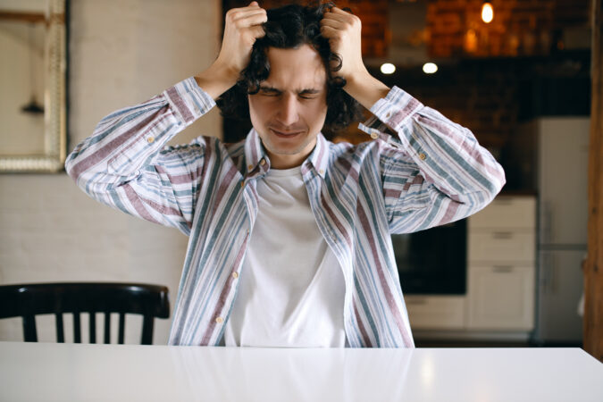 Young man with dark curly hair sitting at white table next to black chair pulling his hair with both hands.