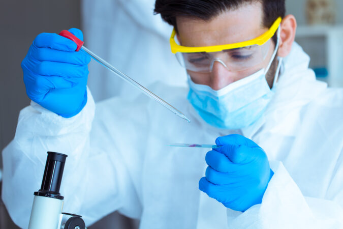 Gowned scientist in goggles, mask, and gloves holding a slide and pipette. Top of microscope visible.